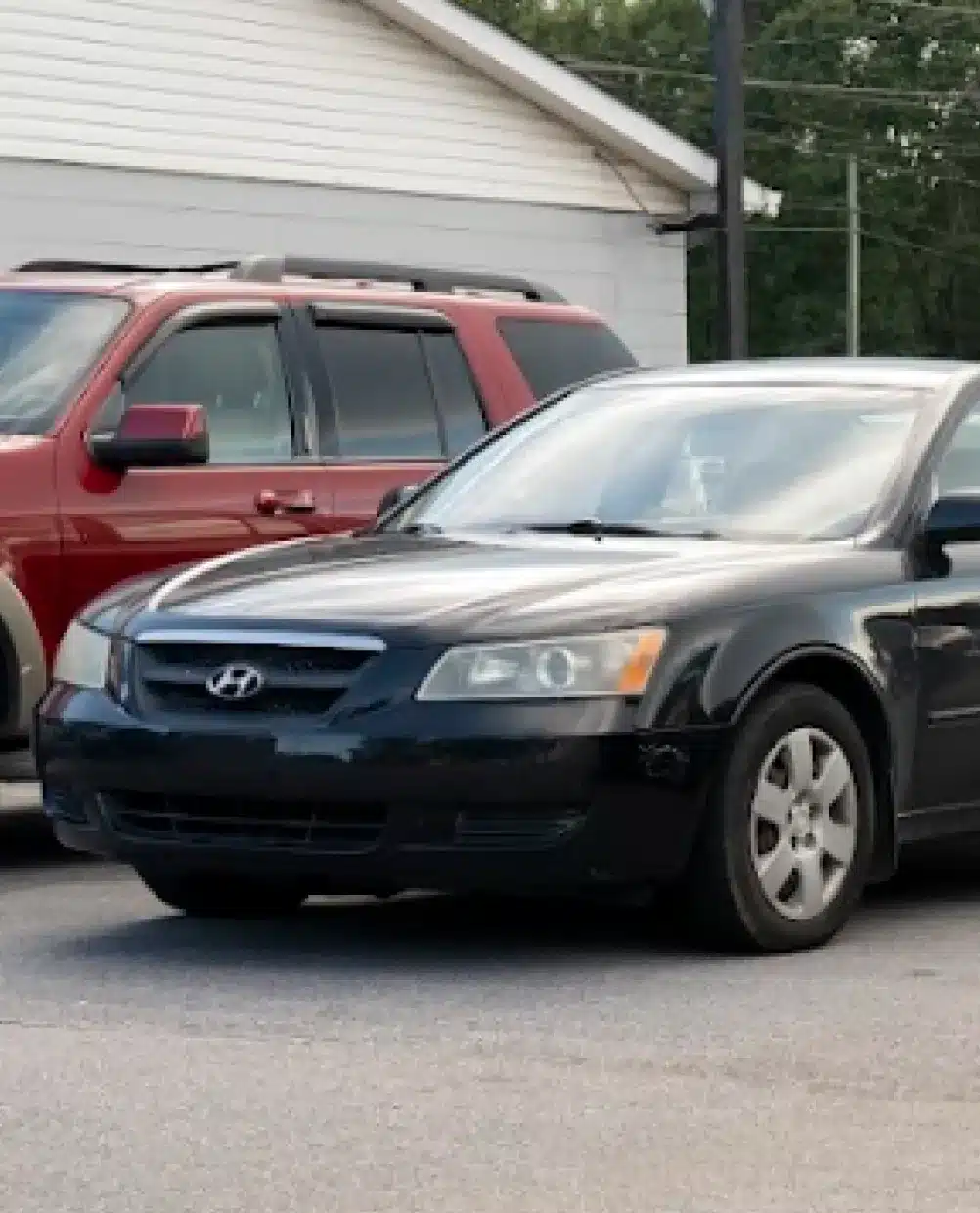 an image showing Asian vehicle repair services in Piedmont, SC at Palmetto Complete Auto Care represented by a black Hyundai sedan is parked in front of a red SUV in an outdoor parking lot near a white building.