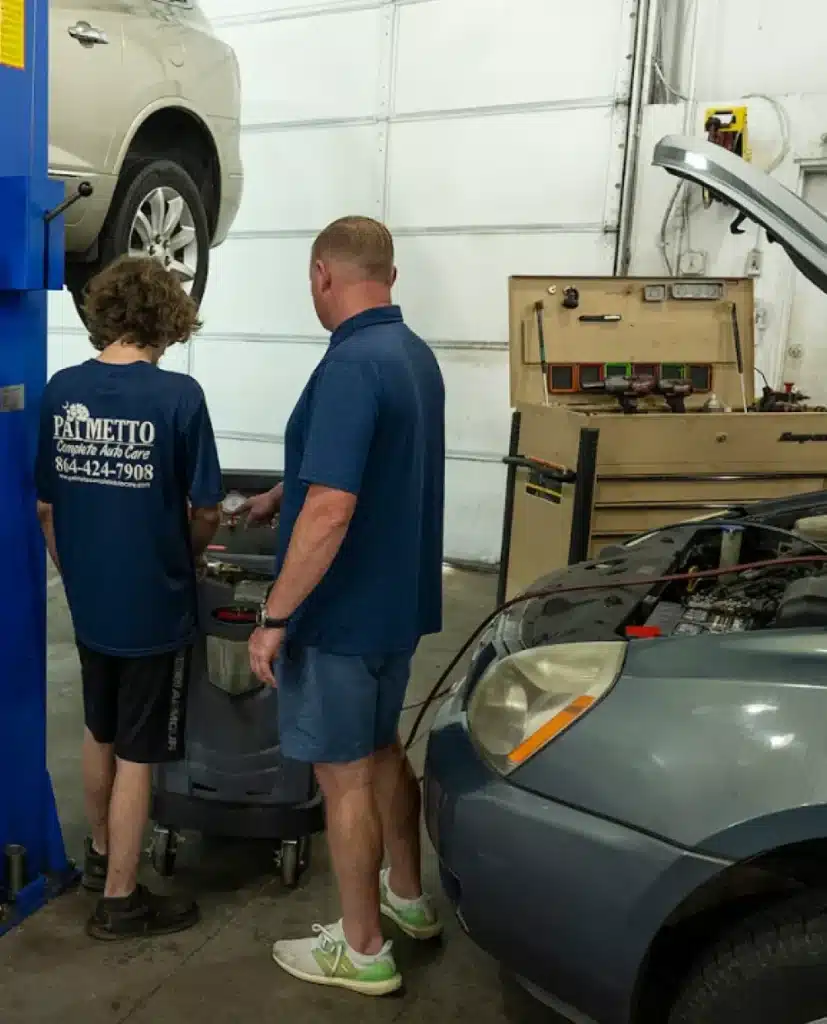 Auto Air Conditioning & Heat Repair by Palmetto Complete Auto Care shown by Two people stand next to a machine in an auto repair shop, examining something together. A car is lifted on a hydraulic lift, and another car with its hood open is nearby. Tools and equipment are visible in the background.