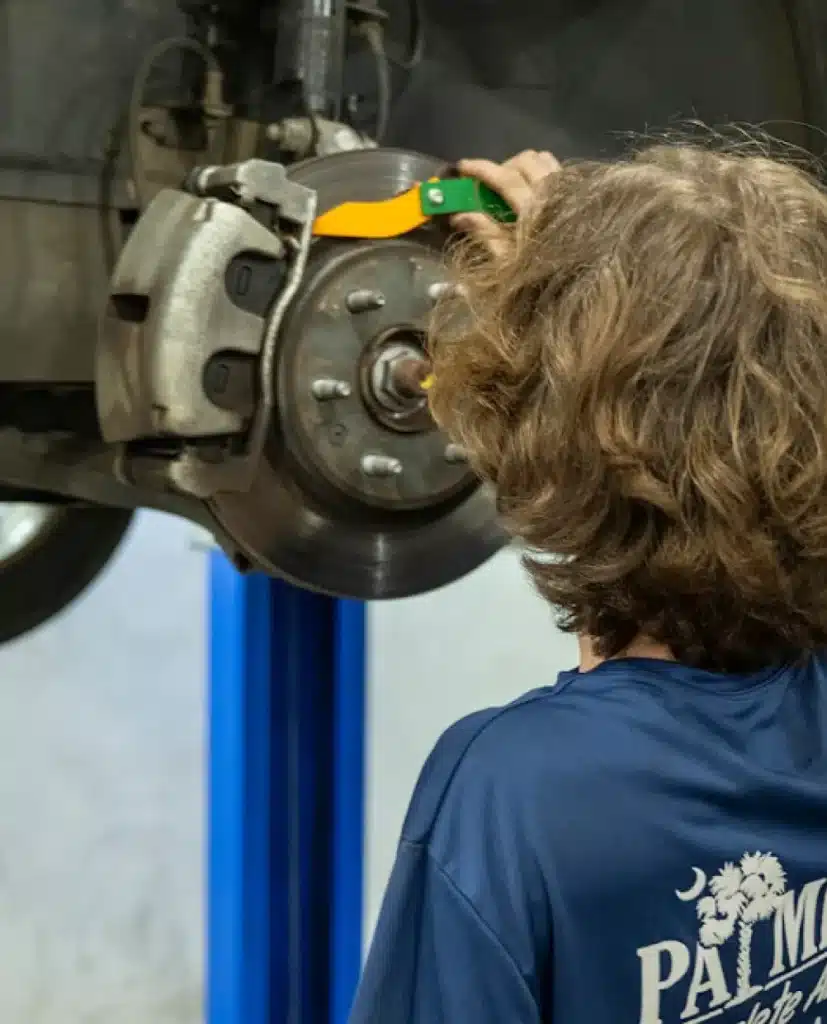 Brake repair being performed in Palmetto Complete Auto Care using a tool to work on the brake system of a car lifted on a blue automotive lift. The person is wearing a blue shirt with partial white text on the back.