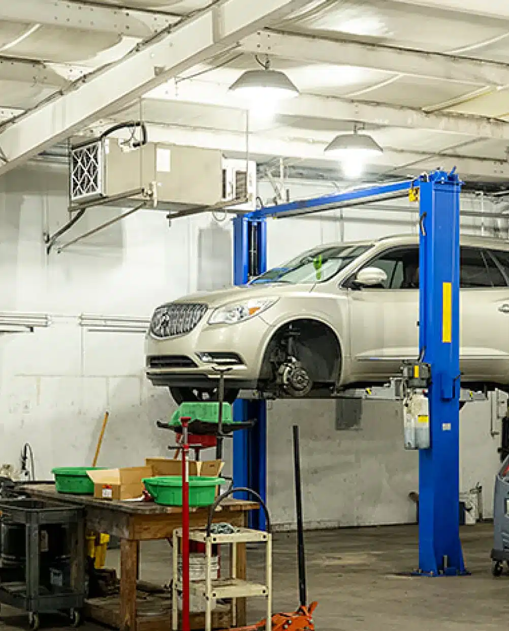 an image showing Domestic vehicle repair services in Piedmont, SC at Palmetto Complete Auto Care with a silver car is lifted on a blue hydraulic car lift in an auto repair shop, with one front wheel removed. Tools and equipment are scattered on workbenches nearby. The shop has white walls and overhead lighting.