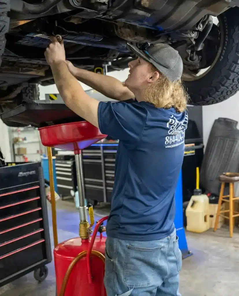 A mechanic with long hair, wearing a cap and blue shirt, works under a raised vehicle in a garage, using tools and draining oil into a red container.