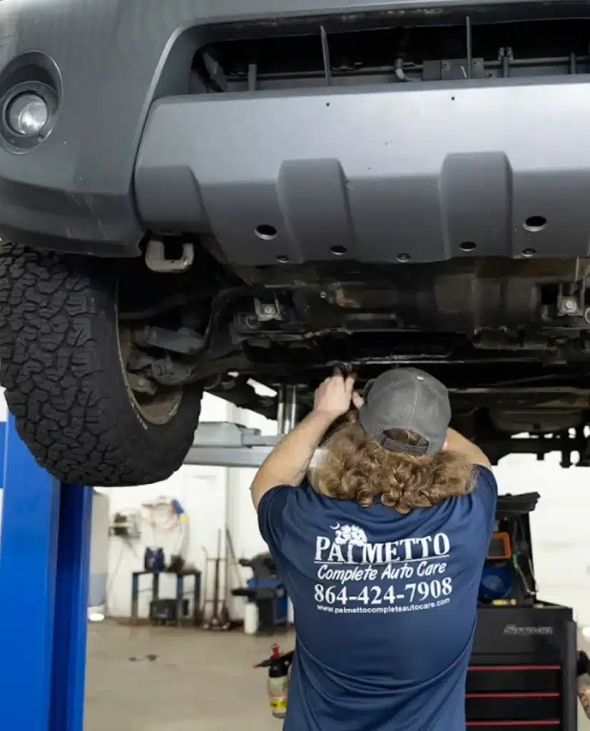 Mechanic in blue Palmetto Complete Auto Care shirt performing suspension repair under a raised vehicle in Piedmont, SC auto repair shop