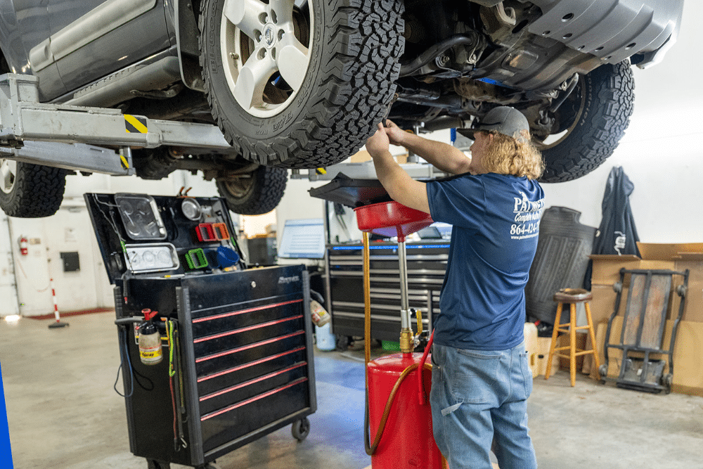 Preventative Maintenance. Auto repair in Piedmont, SC. Palmetto Complete Auto Care. A mechanic performs maintenance under a lifted SUV inside an auto repair shop, using a red oil drain container and surrounded by a tool chest and workshop equipment.